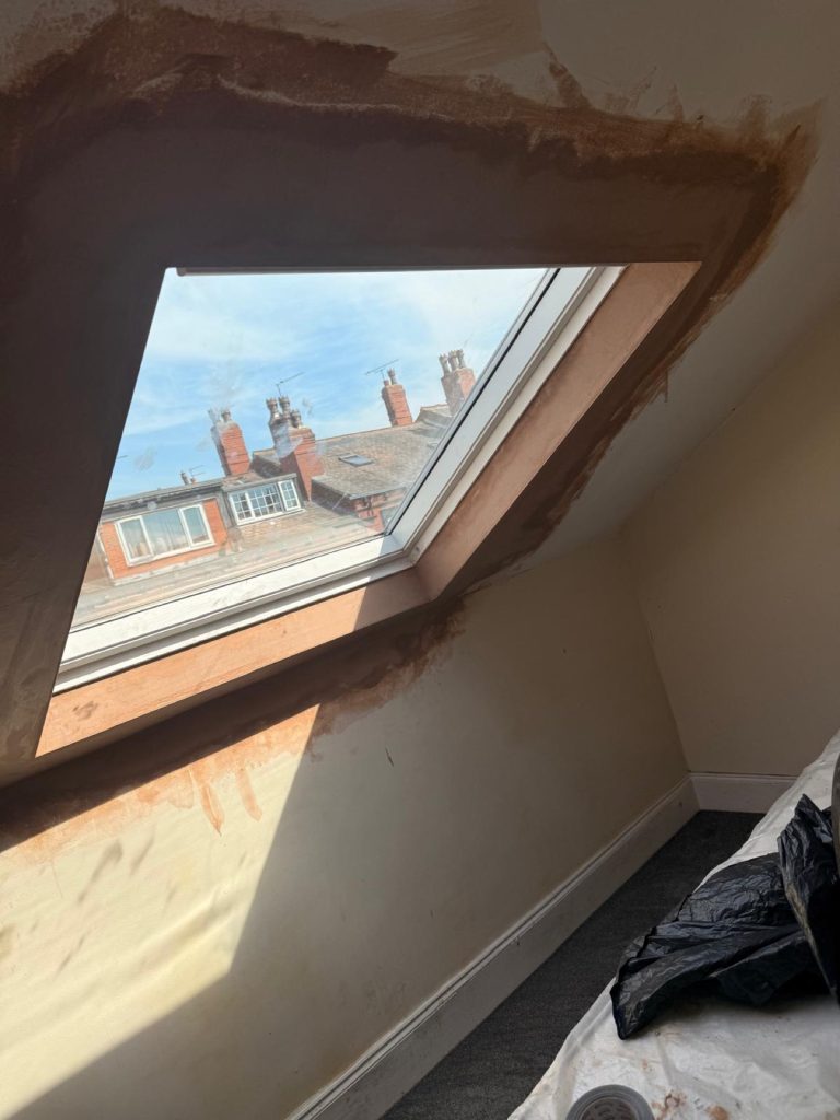 Loft interior with a fresh Velux skylight, plaster patched and drying around the reveal, view of Leeds chimneys outside.